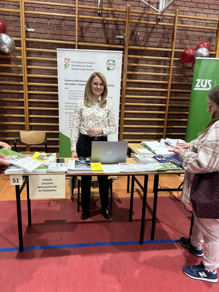 Pokaż zdjęcie: Stanowisko informacyjne Centrum Informacyjno-Doradczego PFRON na Targach Pracy w Szamotułach. Przy stole z rozłożonymi materiałami edukacyjnymi, broszurami i otwartym laptopem stoi kobieta w jasnej, wzorzystej koszuli. W tle widoczny jest baner informacyjny instytucji z logotypem PFRON.