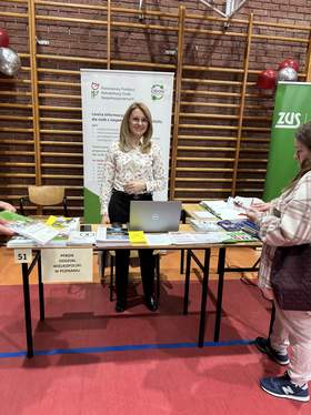 Stanowisko informacyjne Centrum Informacyjno-Doradczego PFRON na Targach Pracy w Szamotułach. Przy stole z rozłożonymi materiałami edukacyjnymi, broszurami i otwartym laptopem stoi kobieta w jasnej, wzorzystej koszuli. W tle widoczny jest baner informacyjny instytucji z logotypem PFRON.