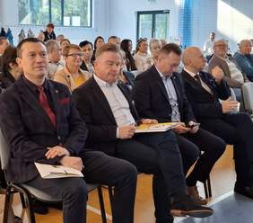 Pokaż zdjęcie: Na krzesłach ułożonych w rzędach siedzą ludzie. Pięć mężczyzn w garniturach siedzi w pierwszym rzędzie na krzesłach, słuchając przemówienia.