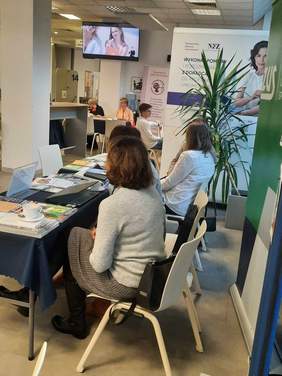 Pokaż zdjęcie: W Sali Obsługi Klientów Łódzkiego Oddziału NFZ znajdują się stoliki, przy których siedzą różne osoby. Na stolikach leżą materiały promocyjne i reklamowe różnych instytucji. 