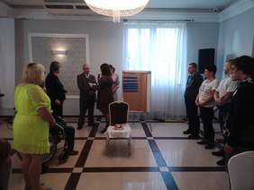 Pokaż zdjęcie: Po prawej i lewej stronie na scenie stoi po kilka osób biorących udział w konkursie na temat Grecji. Kobieta z mikrofonem podchodzi do uczestników i zadaje im pytania na temat Grecji