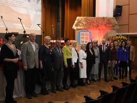 Pokaż zdjęcie: Na scenie, w rzędzie stoi grupa 14 osób, pozują do pamiątkowego zdjęcia.