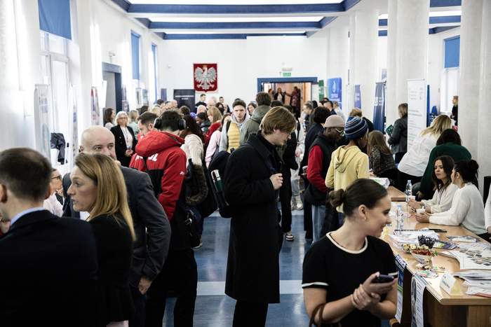 Pokaż zdjęcie: Korytarz z rzędami stolików przy których rozmawiają wystawcy oraz uczestnicy wydarzenia., Na stolikach leżą ulotki, foldery gratisy.