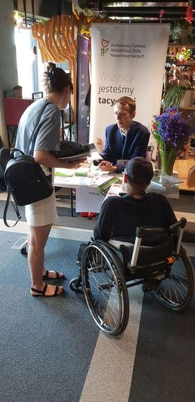 Pokaż zdjęcie: Mężczyzna przy stoisku Oddziału Wielkopolskiego PFRON udziela informacji młodej kobiecie wraz z mężczyzną poruszającym się na wózku. Na stoisku ułożone zostały materiały informacyjne oraz ustawione dwa bukiety kwiatów. W tle widoczny jest baner PFRON.