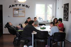 Spotanie przy stole, jedna osoba na wózku inwalidzkim w postawie półleżącej. Obok tablica z napisem "Aktywni"