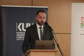 Pokaż zdjęcie: Przy mównicy stoi mężczyzna z brodą ubrany w ciemny garnitur