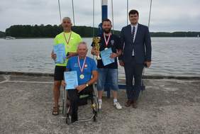 Pokaż zdjęcie: 4 mężczyzn na tle jeziora, 3 stoi, a jeden siedzi na wózku. Trzech z nich ma w rękach dyplomy i medale