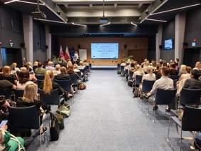 Szerokie ujęcie tej samej sali konferencyjnej z tyłu, przedstawiające liczną publiczność siedzącą w rzędach krzeseł skierowanych stronę sceny. W tym momencie na scenie przy mównicy stoi kobieta, a na dużym ekranie w tle wyświetlana jest jasna prezentacja z wypunktowaną treścią. Po lewej stronie sceny stoją flagi państwowe i unijne. Wnętrze sali ma ciemne ściany i nowoczesne, liniowe oświetlenie sufitowe.