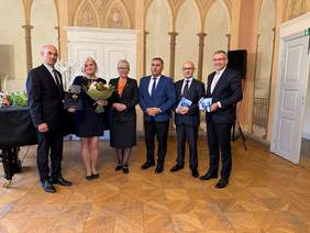 Pokaż zdjęcie: W Sali muzealnej z namalowanymi łukami na ścianach, stoją dwie kobiety i czterej mężczyźni. Wszyscy ubrani na galowo. Jedna z nich trzyma w ręku kwiaty. Mężczyzna obok medal w granatowym pudełku.