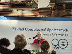 Pokaż zdjęcie: Na zdjęciu znajduje się kobieta w średnim wieku o blond włosach, posiadająca okulary, elegancko ubrana w czerwona garsonkę. Przemawia do mikrofonu, stojąc za przezroczystą mównicą. W tle widoczny jest baner z napisem "Zakład Ubezpieczeń Społecznych" oraz hasłem związanym z obchodami 90-lecia instytucji, oznaczonymi datami "1934–2024". W górnej części kadru można dostrzec elementy dekoracyjne wnętrza, takie jak ozdobne drewniane detale.  Atmosfera jest oficjalna i poważna, typowa dla uroczystych spotkań i jubileuszów.