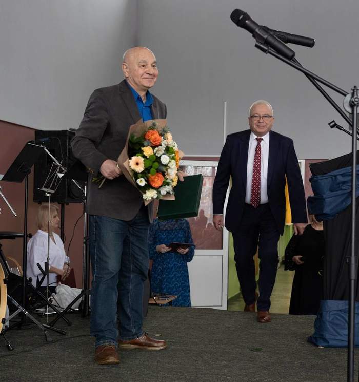 Pokaż zdjęcie: Na scenie przy mikrofonie stoi mężczyzna w garniturze, w ręku trzyma kwiaty.