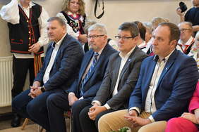 Pokaż zdjęcie: Na widowni w pierwszym rzędzie siedzi czterech mężczyzn. Wszyscy są ubrani w garnitury. W tle widać członków zespołów ludowych. 