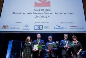Pokaż zdjęcie: Dwie kobiety i trzech mężczyzn stoją na scenie. Wszyscy ubrani na galowo. Mężczyźni trzymają w rękach dyplomy i kwiaty. Nad nimi telebim z napisem: Gala 30-lecia Stowarzyszenia Pomocy Osobom Autystycznym Żyć Razem.