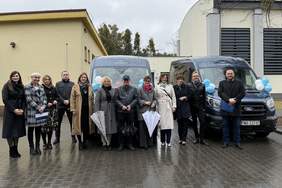 Pokaż zdjęcie: Zdjęcie grupowe kilkunastu osób stojących w jednym rzędzie przed dwoma ciemnoniebieskimi busami udekorowanymi biało-niebieskimi balonami. Uczestnicy wydarzenia ubrani są w płaszcze i kurtki, część z nich trzyma parasole. Wszyscy stoją na zewnątrz na mokrej kostce brukowej.