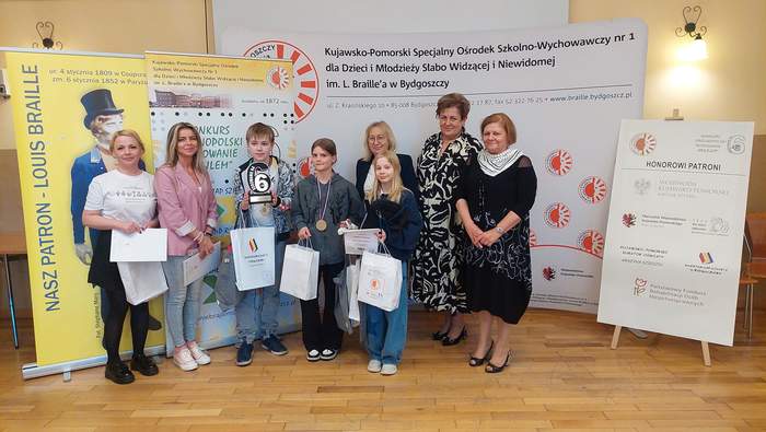 Od lewej stoją uczestnicy konkursu - laureaci i trzymają w rękach dyplomy i torebki upominkowe. Od prawej strony stoją Dyrektor Kujawsko-Pomorskiego Oddziału PFRON Aleksandra Gierej oraz Kujawsko-Pomorski Wicekurator Oświaty Joanna Rec. W tle widać banery.