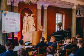 Pokaż zdjęcie: Sala konferencyjna z publicznością siedzącą na krzesłach i zwróconą w stronę sceny. Na środku widać mównicę, za którą stoi osoba prowadząca prezentację. Po lewej stronie sceny ustawiony jest ekran projekcyjny i wyświetlony jest na nim slajd. Za mównicą znajdują się wysokie kolumny oraz duża jasna rzeźba przedstawiająca dwie stojące postacie ludzkie na postumencie.  Ściany mają ciepły, czerwony kolor, a oświetlenie w odcieniach pomarańczowo niebieskich podkreśla kolumny i rzeźbę.  Tekst na ekranie projekcyjnym: „Różne mózgi, jedna klasa”.