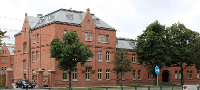 Pokaż zdjęcie: Zabytkowy budynek z czerwonej cegły stojący wzdłuż ulicy. Po prawej bujne zielone drzewa. Po lewej brama z której wyjeżdża ciemny samochód.