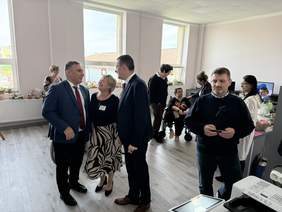 Pokaż zdjęcie: Grupa osób rozmawiających w jasnym pomieszczeniu z dużymi oknami; w tle inni uczestnicy wydarzenia przy stołach z dekoracjami i materiałami. Atmosfera swobodna, część osób zaangażowana w rozmowy.