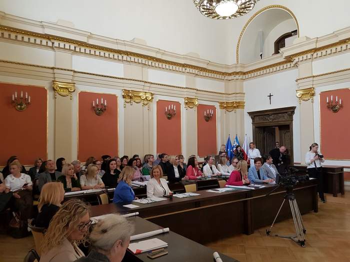 Pokaż zdjęcie: Kilkadziesiąt osób siedzi w sali konferencyjnej, w zabytkowym budynku, w którym można dostrzec wiele różnych elementów zdobiących ściany pomieszczenia. Po środku sali ustawiona została kamera telewizyjna.