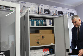 Pokaż zdjęcie: Otwarta szafa wypełniona pudełkami ze sprzętem, przy której stoi mężczyzna w garniturze i prezentuje sprzęt