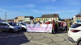 Na miejskim placu w Poddębicach stoi duża grupa osób. Trzymają przed sobą transparent z napisem: Marsz świadomości i akceptacji autyzmu. W tle widać domy i samochody.  