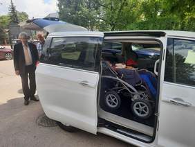 Pokaż zdjęcie: We wnętrzu auta znajduje się osoba siedząca na wózku inwalidzkim. Za samochodem stoi dwóch mężczyzn. Pojazd jest w białym kolorze. 