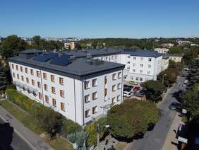 Pokaż zdjęcie: Zdjęcie z lotu ptaka, domu pomocy społecznej, wysokie białe zabudowania pomiędzy drzewami. Zabudowania w kształcie litery U, budynek trzy kondygnacyjny