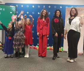 Pokaż zdjęcie: Grupa dzieci i dorosłych stoi na scenie przed niebieskim tłem z logotypami. Kilkoro dzieci w kolorowych, eleganckich strojach unosi ręce i uśmiecha się. Obok nich stoją trzy dorosłe kobiety, również uśmiechnięte, ubrane elegancko.