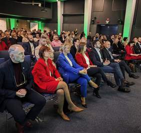 Pokaż zdjęcie: Zdjęcie przedstawia widownię podczas konferencji lub spotkania. Ludzie siedzą w rzędach krzeseł, wielu z nich jest w strojach formalnych lub półformalnych. Na pierwszym planie widoczni są, od lewej, mężczyzna z brodą i okularami trzymanymi w dłoni, kobieta w jaskrawoczerwonej marynarce, kobieta w jasnoniebieskim garniturze, kobieta w czerwonej marynarce oraz dwaj mężczyźni w garniturach. Sala jest oświetlona zielonym światłem, a na suficie widoczne są nowoczesne, linearne lampy.