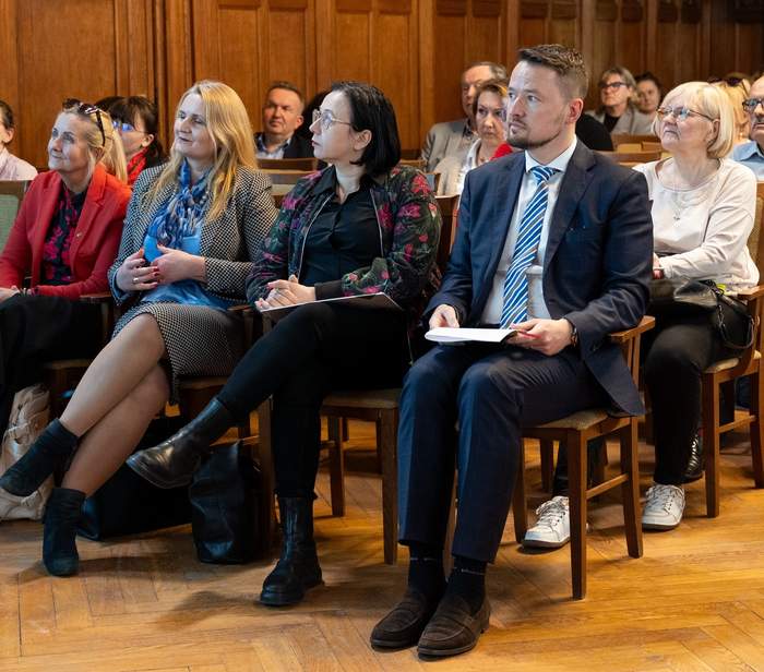 Pokaż zdjęcie: Osoby siedzące na krzesłach ustawionych w rzędzie w sali stylizowanej na dawną, z drewnianymi wykończeniami i zdobieniami. 