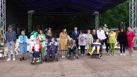 na scenie w plenerze grupa kilkunastu osób, mężczyzn i kobiet w tym 4 osoby na wózkach inwalidzkich. Stoją w rzędzie.