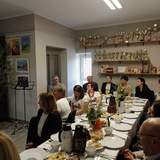 Grupa osób przy długim, zastawionym stole podczas śniadania wielkanocnego w Warsztacie Terapii Zajęciowej w Gołańczy. Na stole biała zastawa, ciasta i wiosenne dekoracje. W tle ściana z licznymi pucharami oraz galeria prac plastycznych. Pomieszczenie jest jasne, wypełnione radosną, świąteczną atmosferą.