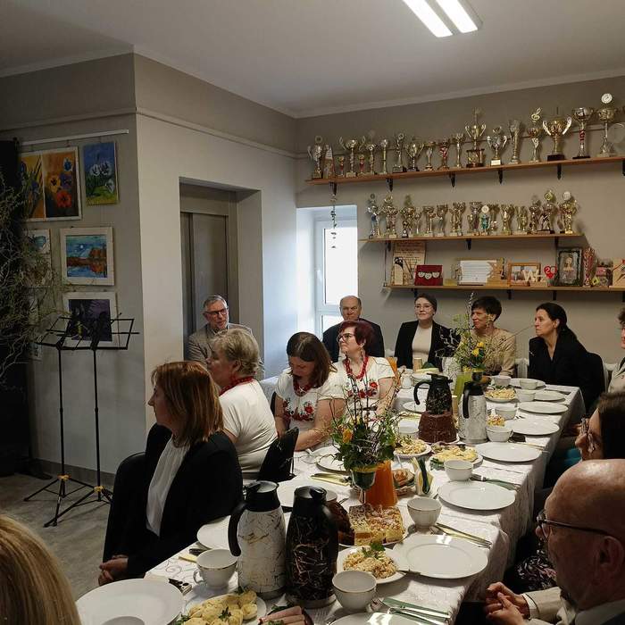 Pokaż zdjęcie: Grupa osób przy długim, zastawionym stole podczas śniadania wielkanocnego w Warsztacie Terapii Zajęciowej w Gołańczy. Na stole biała zastawa, ciasta i wiosenne dekoracje. W tle ściana z licznymi pucharami oraz galeria prac plastycznych. Pomieszczenie jest jasne, wypełnione radosną, świąteczną atmosferą.