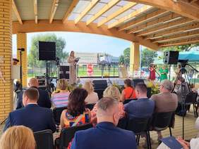 Pokaż zdjęcie: Zdjęcie uczestników uroczystości zrobione z tylnych rzędów, na scenie stoi kobieta z mikrofonem.