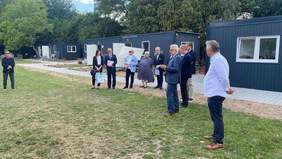 Pokaż zdjęcie: Grupa ludzi stoi w półkolu na trawie przed kontenerami mieszkalnymi, mężczyzna w garniturze przemawia