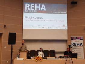 sala konferencyjna, przy stole siedzi kobieta i mężczyzna, za nimi ekran zawierający informacje o wydarzeniu, w szczególności duży napis reha oraz imię i nazwisko prelegenta, z lewej strony za mównicą mężczyzna