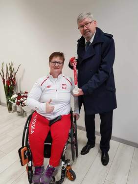 Pokaż zdjęcie: Paraolimpijka siedzi na wózku inwalidzkim i pokazuje kciuk w górę. Obok niej stoi pan w granatowym płaszczu i prezentuje srebrny medal. 