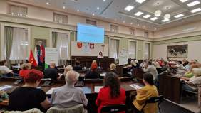 Pokaż zdjęcie: kobieta stoi na mównicy, przed nią słuchacze zasiadający na sali obrad w Urzędzie Miasta w Kielcach