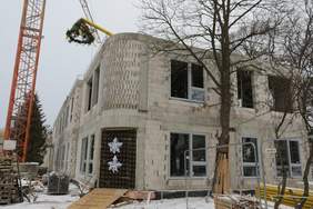 Pokaż zdjęcie: Piętrowy budynek z szarego pustaka z zaokrąglonym narożnym wejściem. Na budynku wisi wiecha wykonana z gałęzi choinki. Do okoła plac budowy, i stojący dźwig. Przed budynkiem dwa drzewa bez liści. Pada śnieg.