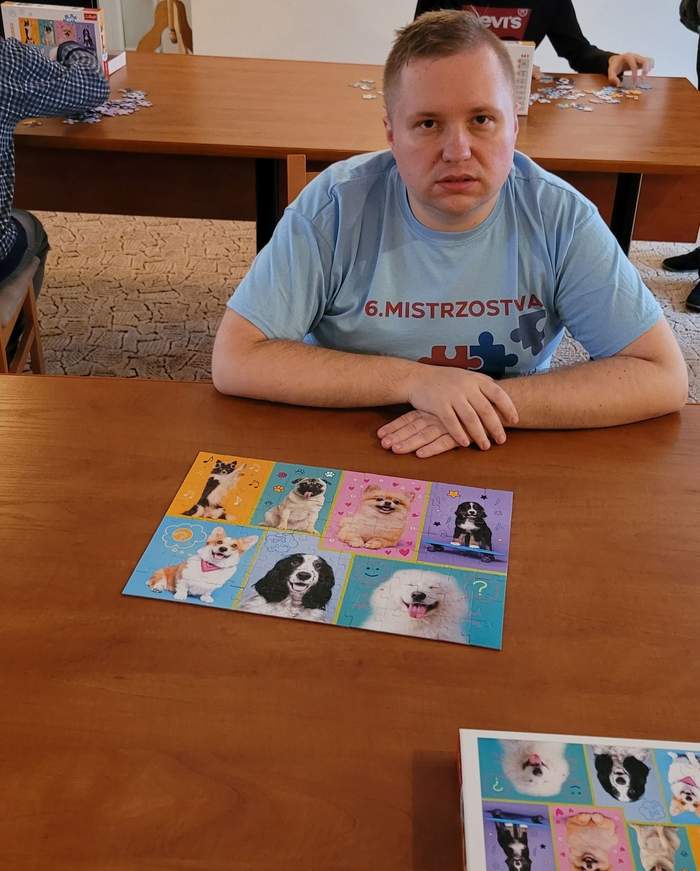 Pokaż zdjęcie: Mężczyzna w niebieskiej koszulce z krótkim rękawem, na której widnieją: napis „6. Mistrzostwa” oraz puzzle w kolorach czerwonym i granatowym, siedzi opierając ręce o duży stół. Na stole widoczny jest ułożony zestaw puzzli prezentujący różne rasy psów, jak również pudełko od tego zestawu. W tle możemy dostrzec także dwie inne osoby układające puzzle, które siedzą przy drugim stole.