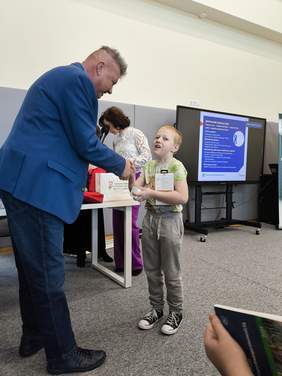 Pokaż zdjęcie: Mężczyzna w garniturze, wręcza nagrodę chłopcu.