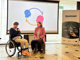 Pokaż zdjęcie: Kobieta na wózku mówi do mikrofonu, obok siedzi druga kobieta z notatnikiem. Za nimi ekran z grafiką dwóch głów połączonych strzałkami. Po prawej roll-up z napisem „Włączamy Pomorskie” i hasłami dotyczącymi integracji społecznej.