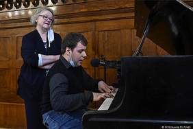 Pokaż zdjęcie: Za fortepianem siedzi mężczyzna ubrany w ciemną koszulę, gra i śpiewa. Z nim stoi kobieta w okularach i ciemnej długiej marynarce. 