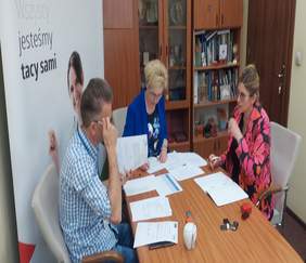 Pokaż zdjęcie: przy stole siedzą trzy osoby, po środku rozłożone dokumenty które są podpisywane po lewej logo PFRON