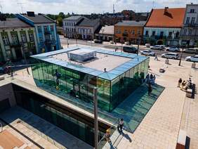 Pokaż zdjęcie: Widok z góry na Stary Rynek w Częstochowie na środku stoi duży przeszklony budynek wokół znajdują się ludzie, samochody i kolorowe kamienice