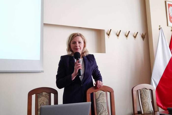 Pokaż zdjęcie: Kobieta blondynka stoi za stołem konferencyjnym, ubrana jest w ciemny garnitur.