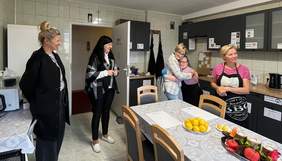 Pokaż zdjęcie: Pięć kobiet w domowej kuchni; dwie stoją przy drzwiach, trzy przy stole, z czego jedna obejmuje drugą. Na stole leżą owoce.