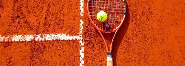 Pokaż zdjęcie: Kawałek ceglastego kortu tenisowego,na nim leży kolorowa rakieta i żółta piłeczka do tenisa.