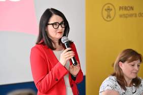 Pokaż zdjęcie: Elegancko ubrana kobieta mówi przez mikrofon. W tle rollup z napisem PFRON wspiera dostępność