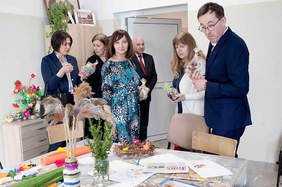 Pokaż zdjęcie: Zdjęcie sali terapeutycznej, przy stole stoi sześć osób, oglądają prace i materiały plastyczne rozłożone na stole.